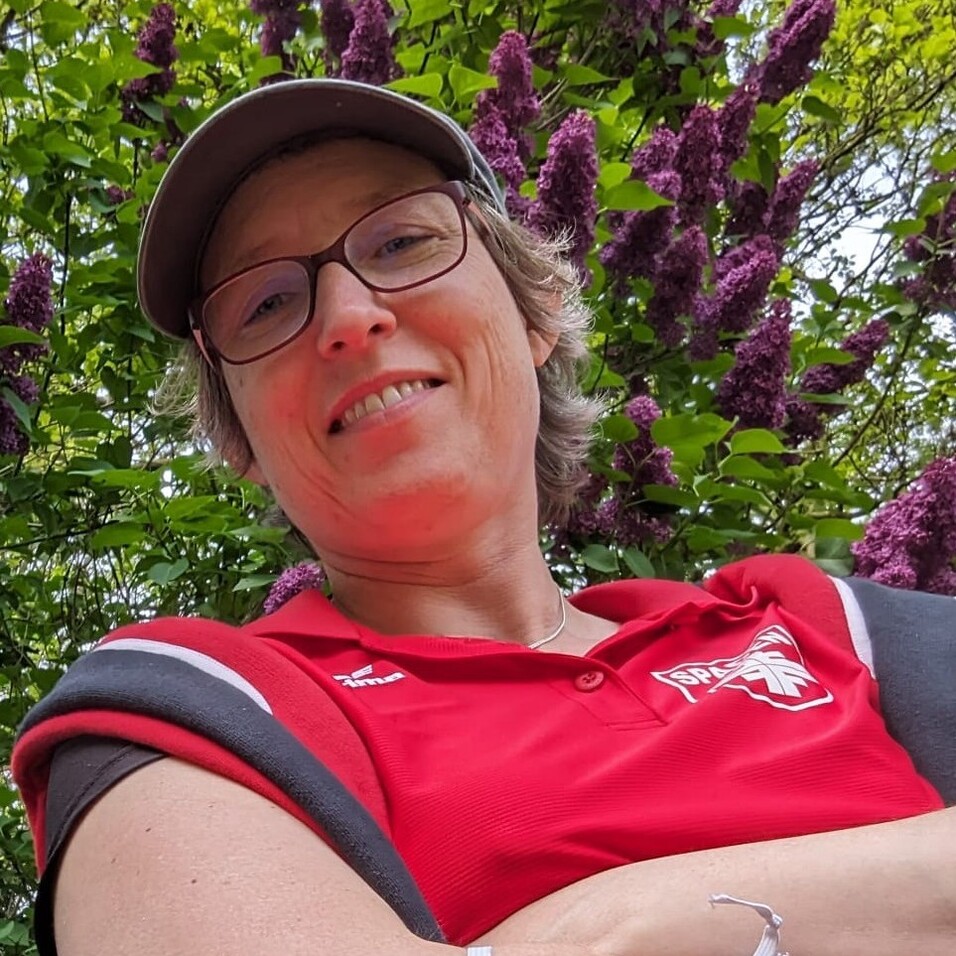 Frau mit Brille und Cap im roten Shirt, umgeben von lila Blumen, sitzt entspannt mit verschr&auml;nkten Armen.