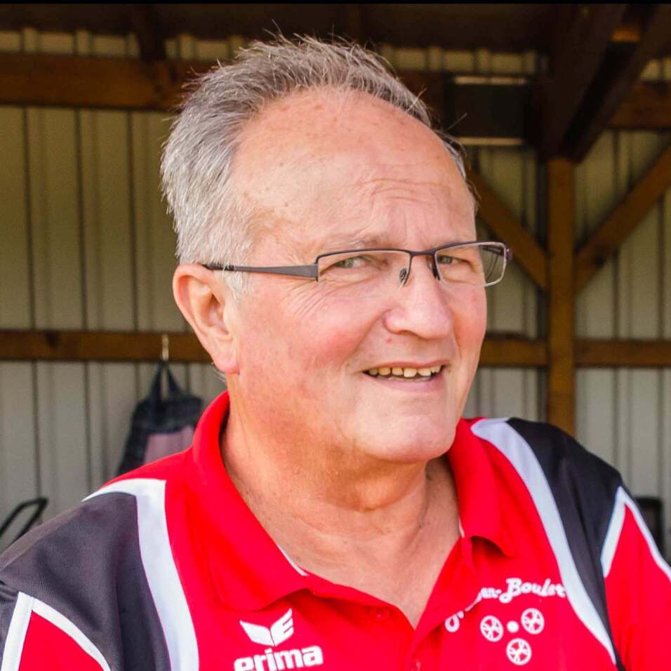 Portrait eines lächelnden Mannes in rotem Sportshirt mit Brille, Stehplatz vor Holzstruktur und neutralem Hintergrund.