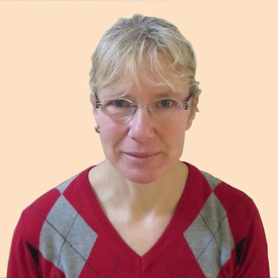 Porträt einer Frau mit kurzen blonden Haaren, Brille und rot-grauer Rautenstrickjacke vor einfarbigem Hintergrund.