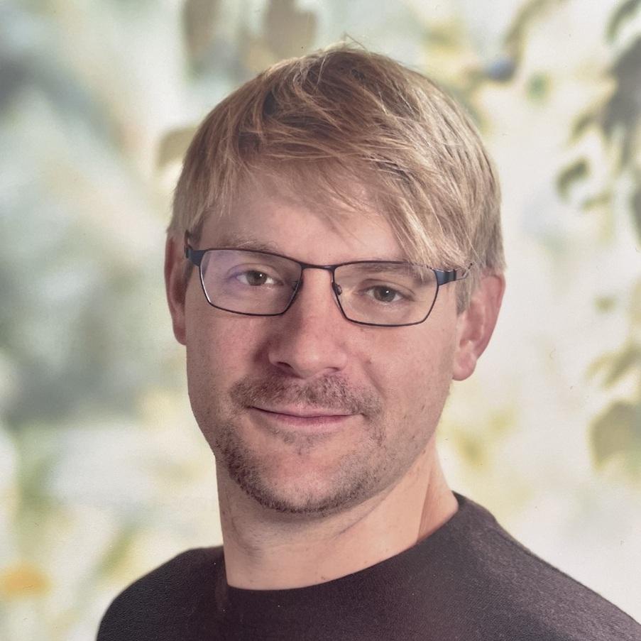 Portrait eines Mannes mit kurzen blonden Haaren und Brille, lächelnd vor einem unscharfen Hintergrund aus Pflanzen.