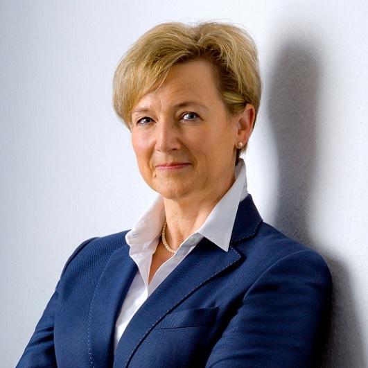 Porträt einer lächelnden Frau mit kurzen blonden Haaren in einem blauen Blazer, an einer Wand stehend.