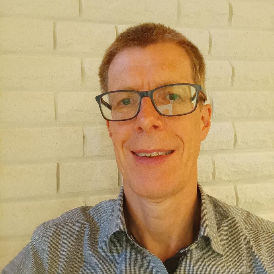 Porträt eines Mannes mit Brille, lächelnd vor einer hellen, strukturierten Wand.