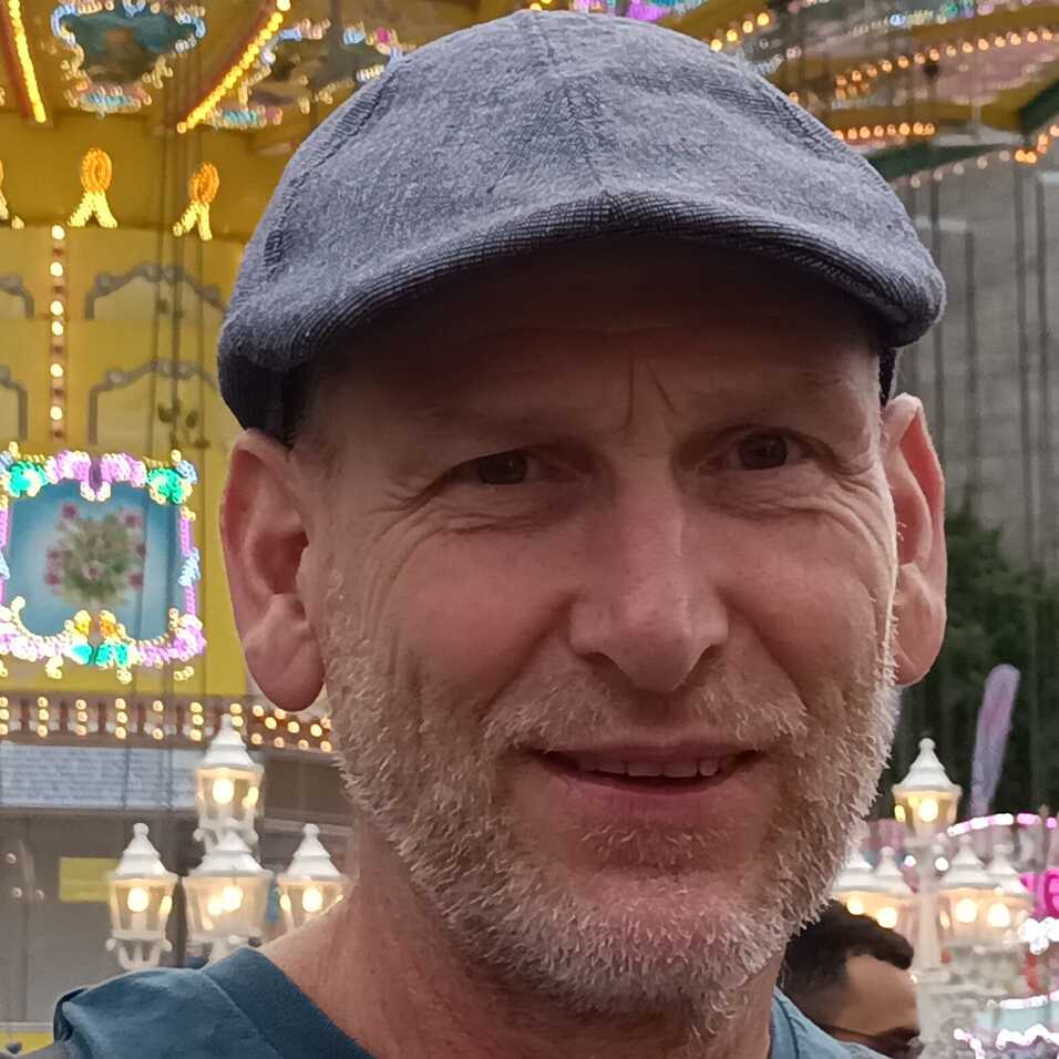 Mann mit grauem Hut lächelt vor einem bunten Jahrmarkt mit nostalgischen Fahrgeschäften.