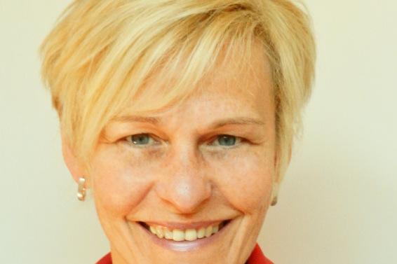Portr&auml;t einer l&auml;chelnden Frau mit blonden Haaren, die ein schwarz-rotes Oberteil tr&auml;gt, vor einem hellen Hintergrund.
