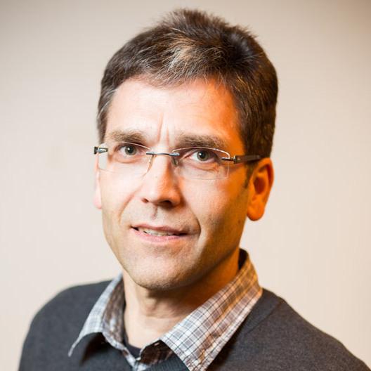 Porträt eines Mannes mit Brille, schütterem Haar und grauem Pullover, lächelt vor einem neutralen Hintergrund.