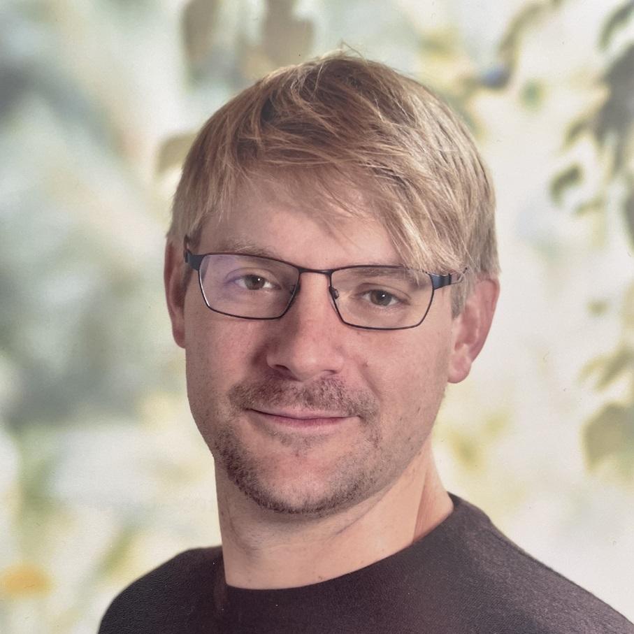 Portrait eines Mannes mit kurzen blonden Haaren und Brille, lächelnd vor einem unscharfen Hintergrund aus Pflanzen.