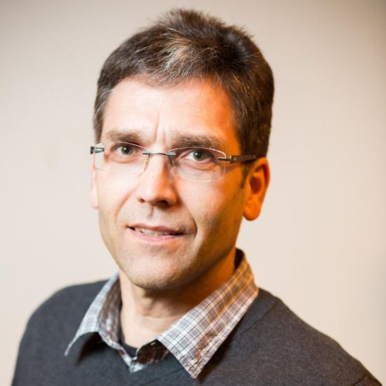 Porträt eines Mannes mit Brille, schütterem Haar und grauem Pullover, lächelt vor einem neutralen Hintergrund.