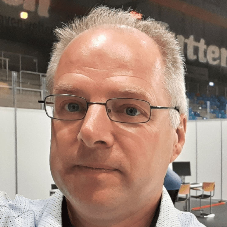 Porträt eines Mannes mit Brille und kurzen, grauen Haaren, in einem Raum mit verstreuten Möbeln im Hintergrund.