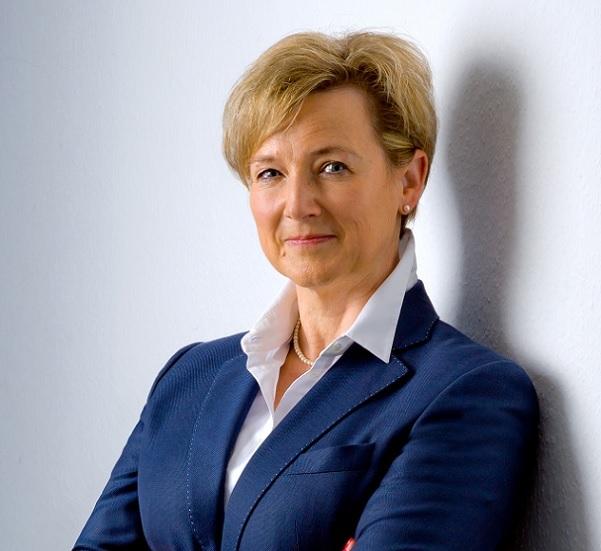 Porträt einer lächelnden Frau mit kurzen blonden Haaren in einem blauen Blazer, an einer Wand stehend.