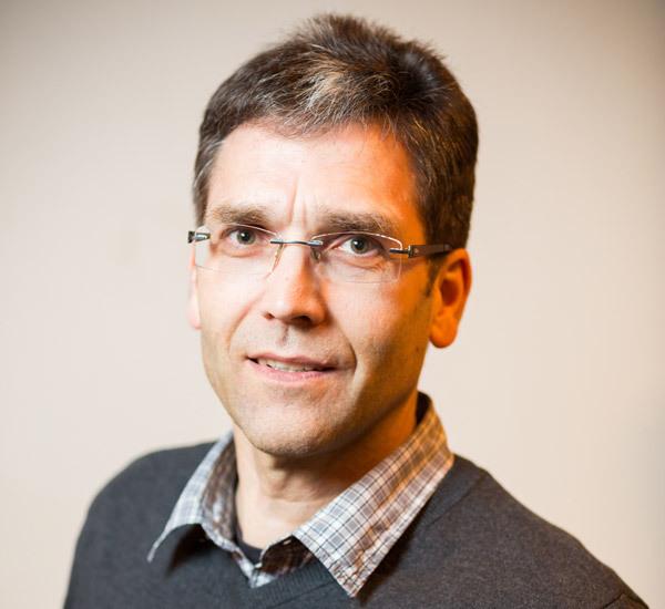 Porträt eines Mannes mit Brille, schütterem Haar und grauem Pullover, lächelt vor einem neutralen Hintergrund.