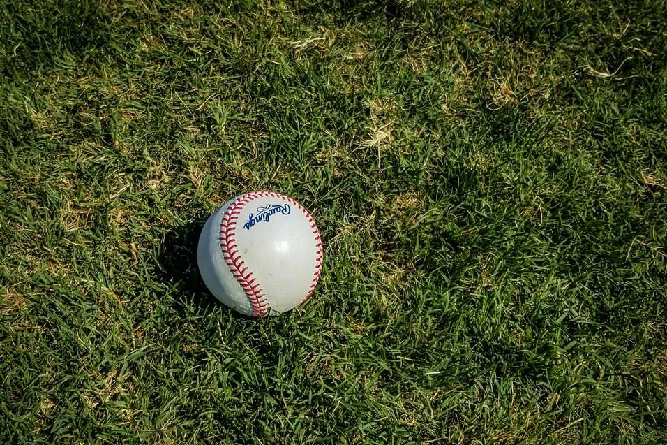 Weißer Baseball mit roter Naht auf grünem Rasen, leicht schräg liegend.