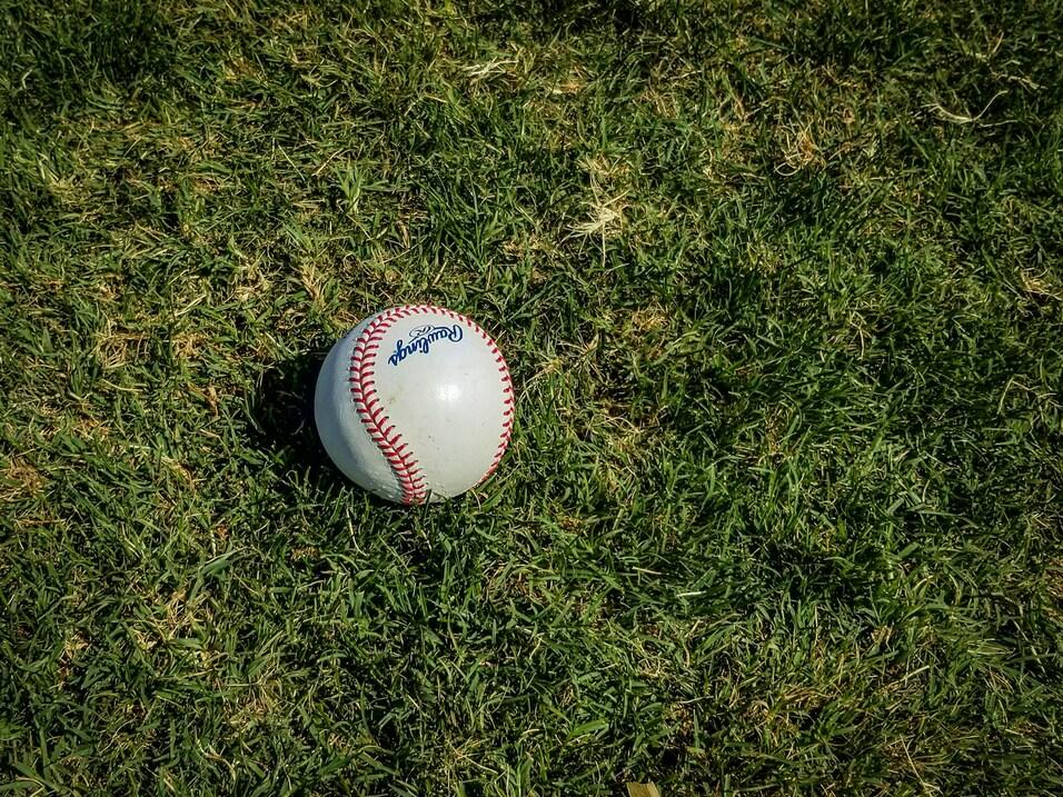 Weißer Baseball mit roter Naht auf grünem Rasen, leicht schräg liegend.