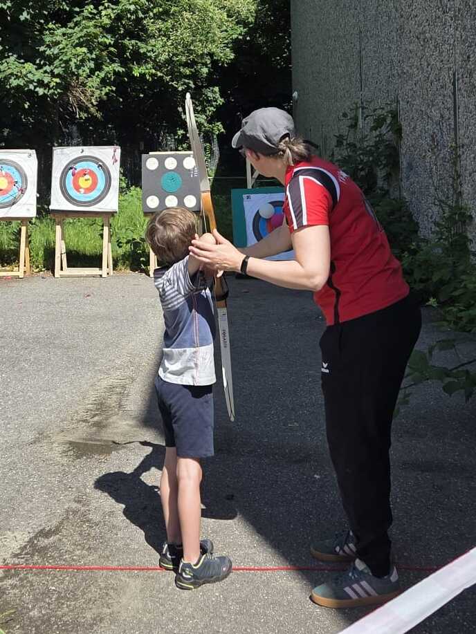 Ein Kind mit einer Pfeil und Bogen-Ausr&uuml;stung wird von einem Trainer angeleitet, w&auml;hrend ein weiterer Erwachsener zuschaut.