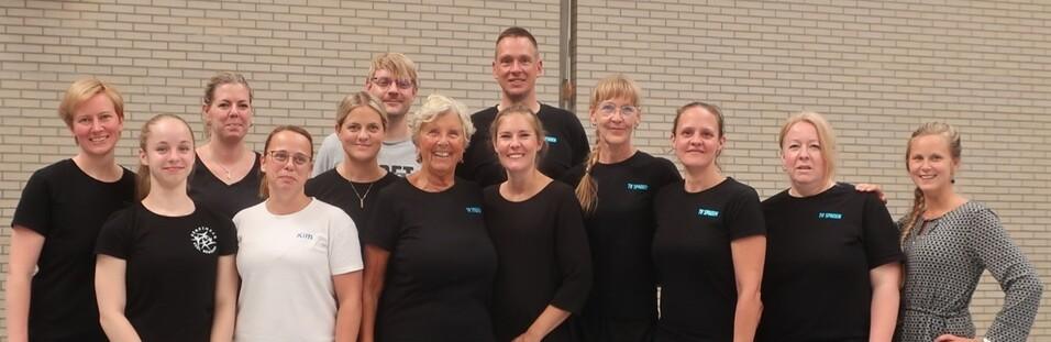 Gruppe von zwölf Personen in sportlicher Kleidung, lächelnd vor einer hellen Wand in einem großen Raum.