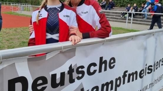 Zwei Frauen in Sportbekleidung stehen an einem Zaun mit dem Banner „Deutsche Mehrkampfmeisterschaften“ im Hintergrund.