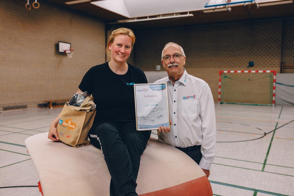 Frau sitzt auf einem Sportgerät, hält ein Zertifikat, neben ihr steht ein Mann mit Brille und lächelt. Sporthalle im Hintergrund.