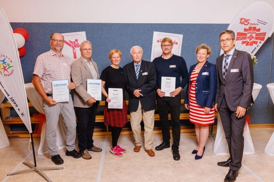 Gruppenfoto von sechs Personen mit Urkunden, vor einem blauen Hintergrund und festlicher Dekoration.