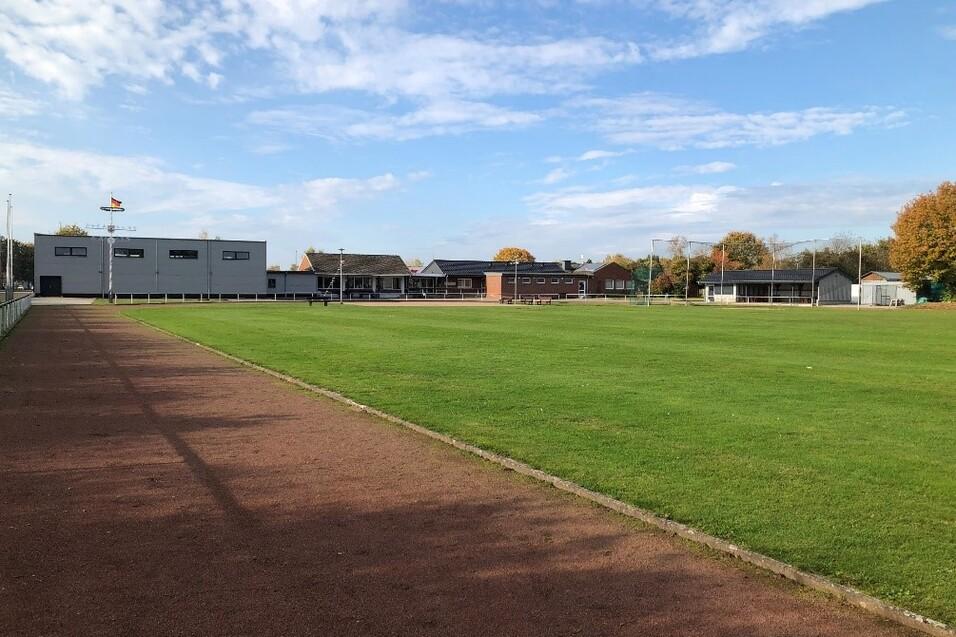 Panorama eines Sportplatzes mit gr&uuml;nem Rasen und mehreren Geb&auml;uden im Hintergrund unter einem blauen Himmel.