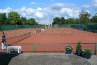 Tennispl&auml;tze mit roten Bel&auml;gen, Umz&auml;unung, Zuschauerb&auml;nken und gr&uuml;ner Umgebung unter einem blauen Himmel mit Wolken.