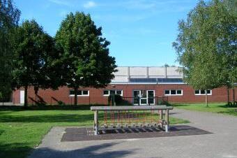 Pflasterweg vor einem roten Geb&auml;ude mit gro&szlig;en Fenstern, umgeben von B&auml;umen und Rasenfl&auml;chen.