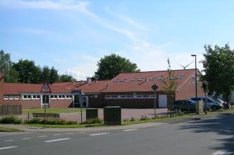 Einfamilienhaus mit roten D&auml;chern, Parkplatz und B&auml;umen an einer ruhigen Stra&szlig;e unter blauen Himmel.