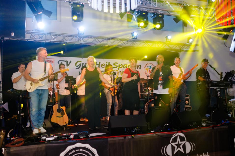 Bandmitglieder stehen auf einer B&uuml;hne mit Instrumenten, umgeben von bunten Lichtern und einer begeisterten Menge.