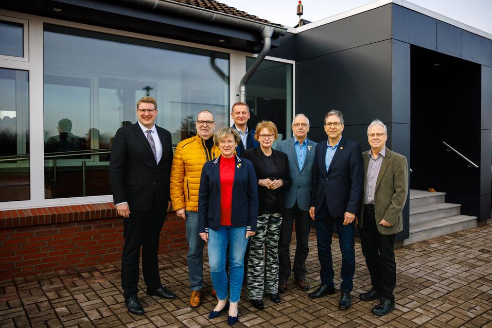 Gruppe von acht Personen in formeller Kleidung, stehend vor einem modernen Geb&auml;ude mit Fenstern und Treppe.