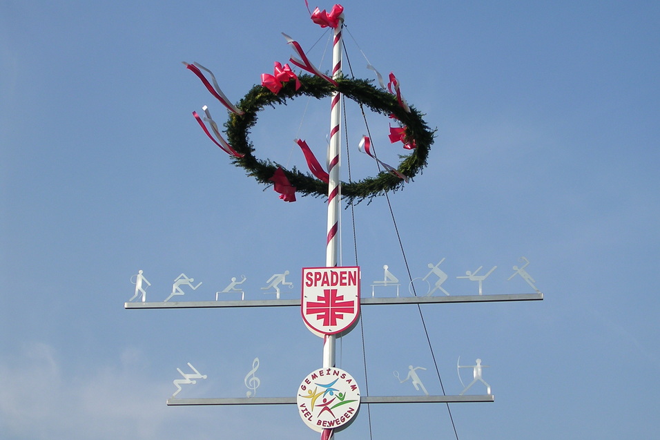 Maibaum mit Kranz, roten Schleifen und Figuren, die verschiedene Sportarten darstellen, vor blauem Himmel.