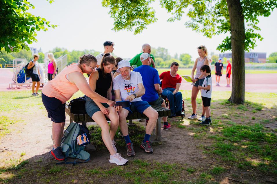 Gruppenszene mit mehreren Personen, die sitzen und ein Tablet betrachten, im Hintergrund Menschen auf einem Sportplatz.