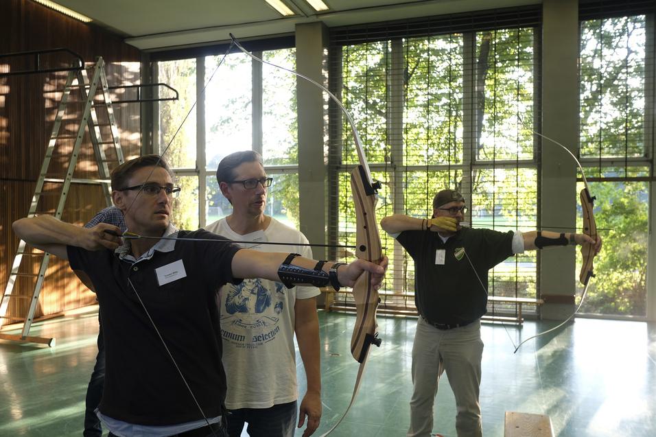 Drei Männer mit Bögen üben das Bogenschießen in einem lichtdurchfluteten Raum mit großen Fenstern.