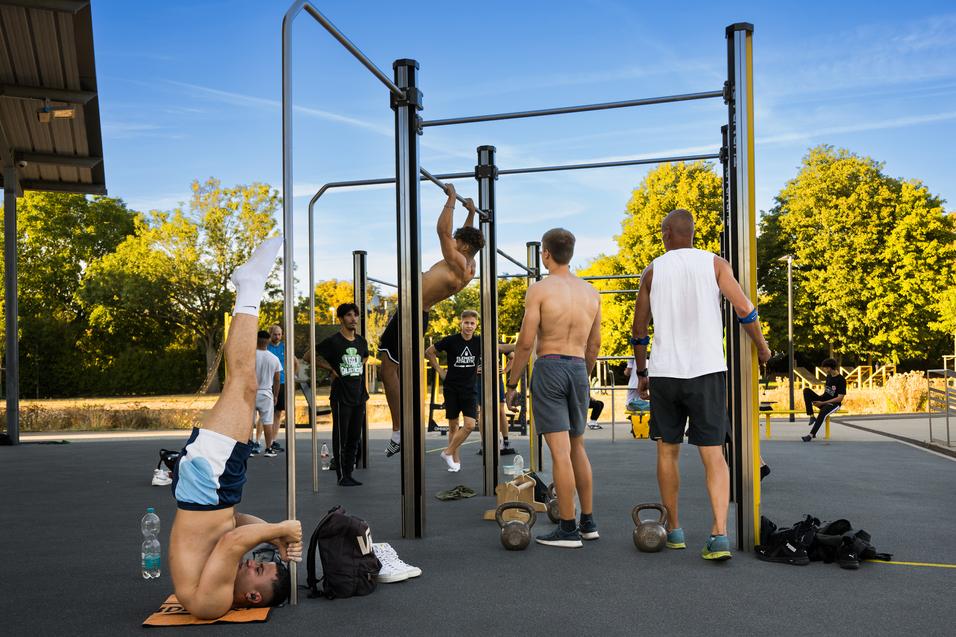 Gruppe von Männern beim Training an Kraftstationen im Freien, einer macht eine Kopfstandübung.