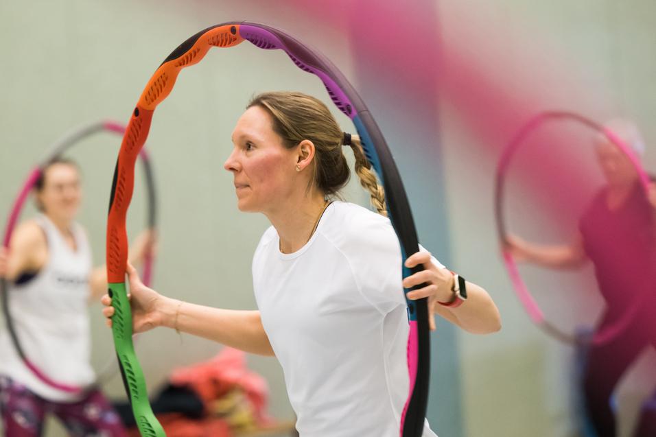 Frau in Sportkleidung hält einen bunten Hula-Hoop-Reifen in einer Turnhalle, im Hintergrund weitere Teilnehmerinnen.