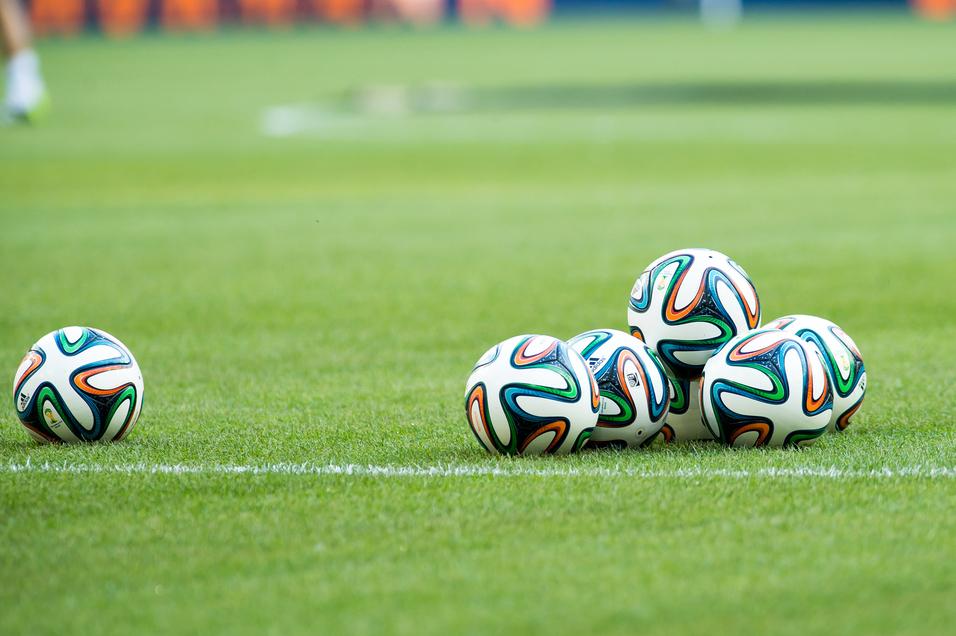 Fünf Fußballbälle liegen auf einem grünen Rasenfeld, sichtbar sind auch der Spielfeldrand und eine verschwommene Hintergrundszene.