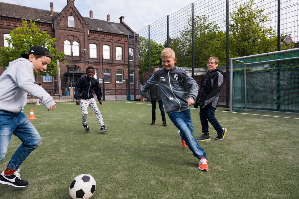 Fünf Kinder spielen Fußball auf einem Kunstrasenplatz, während ein Jugendgebäude im Hintergrund zu sehen ist.