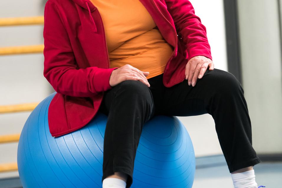 Ältere Frau in roter Jacke sitzt auf einem blauen Gymnastikball in einer Sporthalle.
