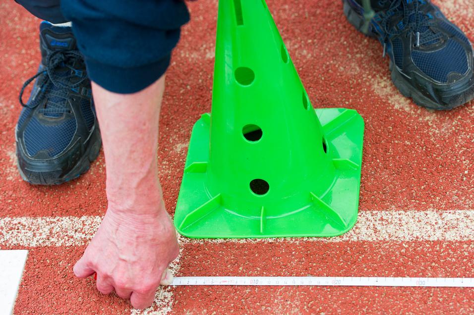 Hand misst mit einem Zollstock die Distanz zu einem grünen Pylonen auf einer roten Sportfläche.