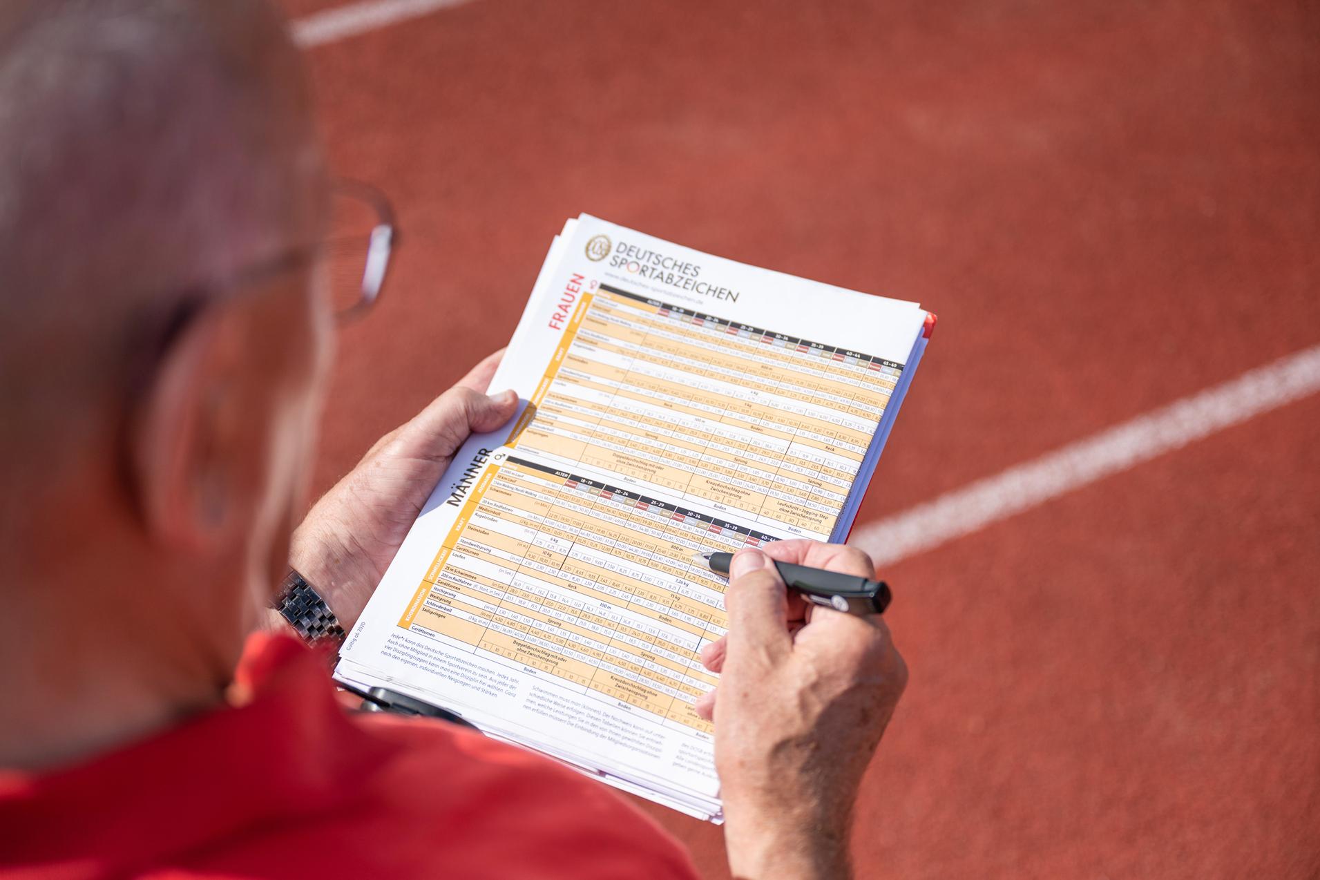 Hand eines Prüfers, der auf einem Blatt mit Sportabzeichen-Anforderungen auf einer Laufbahn notiert.