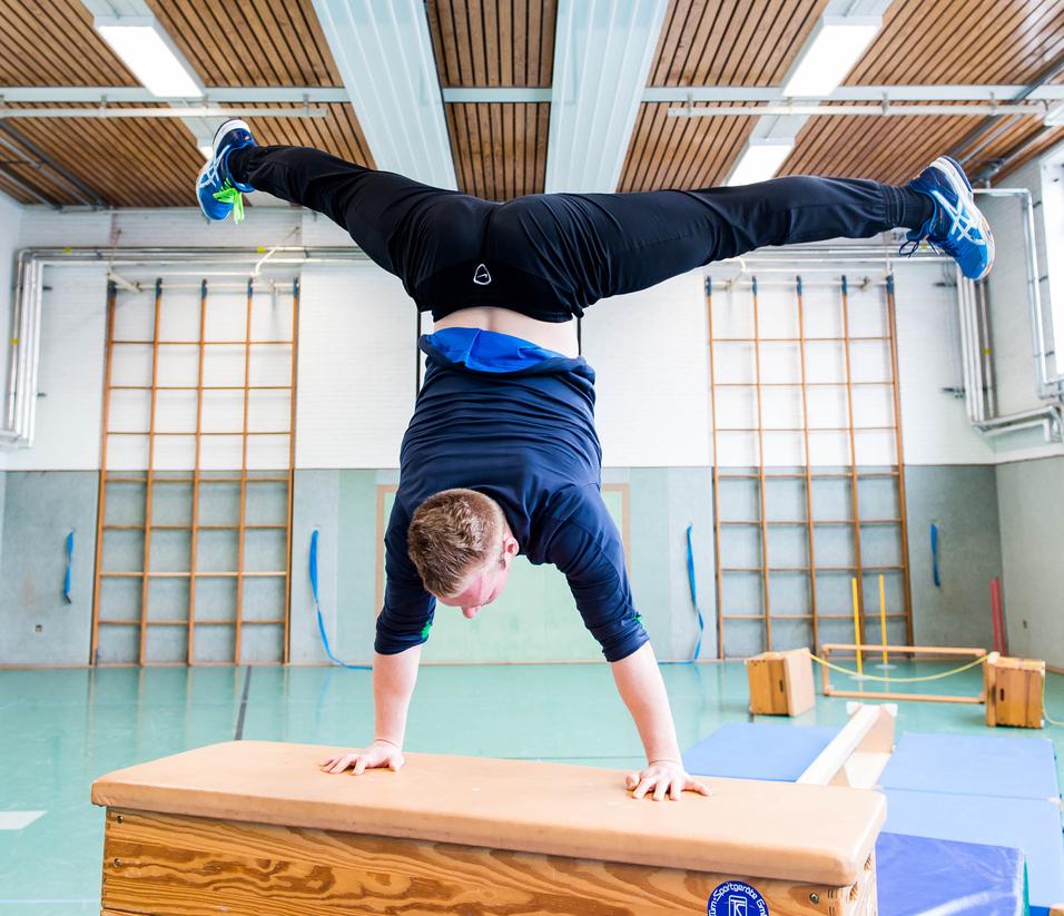 Junge macht Handstand auf einem Holztisch in einer Sporthalle mit verschiedenen Turngeräten im Hintergrund.